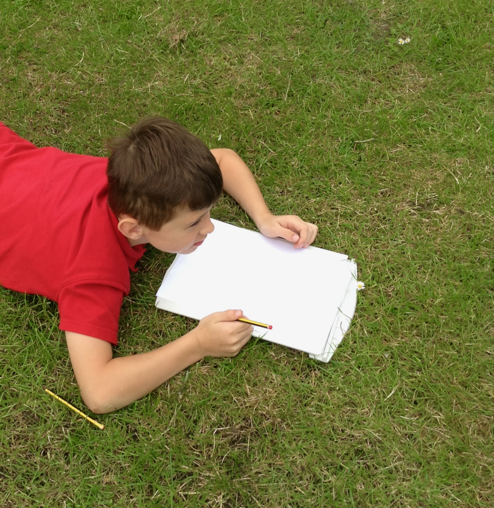 Year Two boy from Hauxton Primary School sitting on grass completing worksheets about trees for Inspire with teacher Pamala Stewart